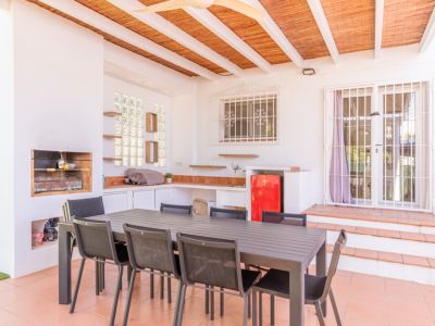 Terraza exterior cubierta con barbacoa integrada, cocina exterior funcional con nevera roja y una amplia mesa de comedor. El espacio cuenta con suelo de terracota y techo de caña.