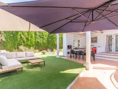 Terraza exterior cubierta con comedor y zona de barbacoa integrada, junto a un césped artificial con mobiliario lounge y una gran sombrilla.