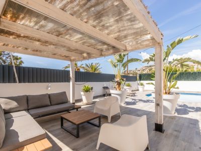 Terraza exterior cubierta con pérgola de madera y techo translúcido, equipada con un sofá modular gris, mesa de centro y plantas tropicales. Suelo de gres y vista parcial a la piscina.