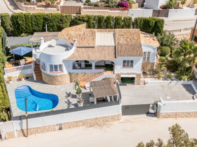 Vista aérea de la villa blanca con tejado de terracota y piscina privada en forma de riñón con mosaico de delfín. Destaca el amplio aparcamiento pavimentado y varias terrazas exteriores.