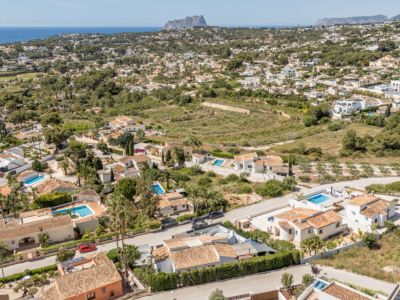 Vista aérea de una zona residencial en Moraira, con múltiples villas de tejados de terracota, piscinas privadas y abundante vegetación. El mar Mediterráneo y un promontorio rocoso lejano enmarcan el paisaje costero.