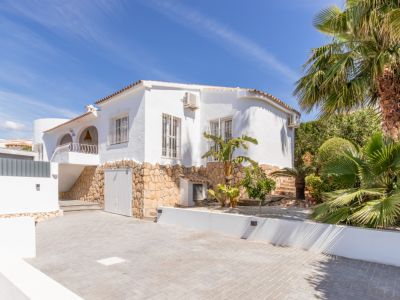 Vista frontal de la villa con fachada blanca, base de piedra natural y tejado de teja. Destaca el amplio parking pavimentado y el exuberante jardín con palmeras.