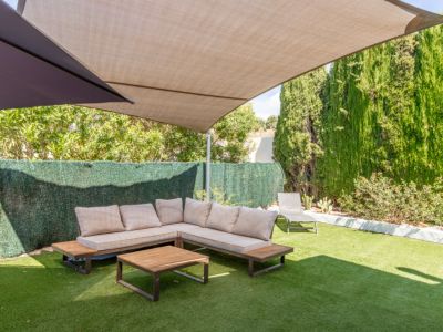 Zona de estar exterior sombreada con sofá en L de cojines claros y mesa de centro de madera sobre césped artificial. Setos altos y árboles ofrecen privacidad bajo una vela de sombra beige.