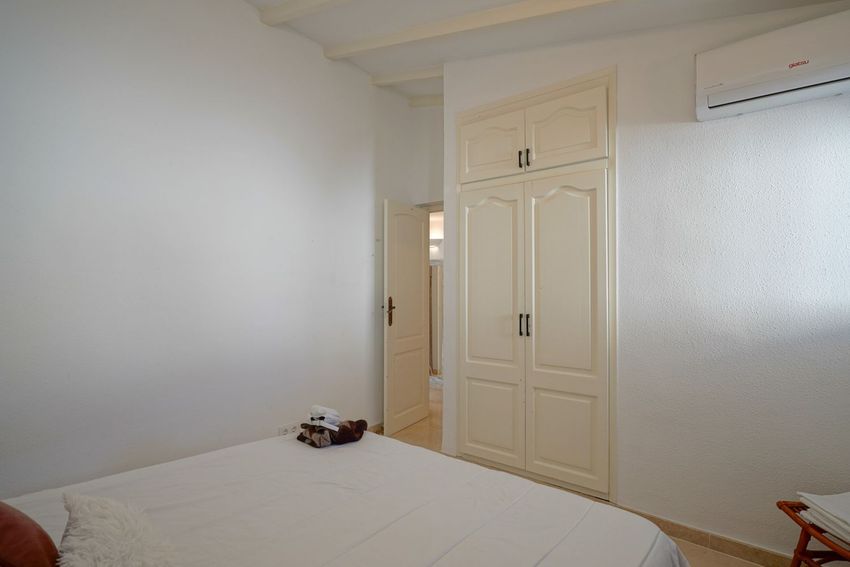 Dormitorio funcional con armario empotrado crema de paneles arqueados y aire acondicionado. Paredes blancas texturizadas, vigas vistas en el techo y suelo de azulejos claros, con puerta abierta. Dormitorio funcional con armario empotrado crema de paneles arqueados y aire acondicionado. Paredes blancas texturizadas, vigas vistas en el techo y suelo de azulejos claros, con puerta abierta.
