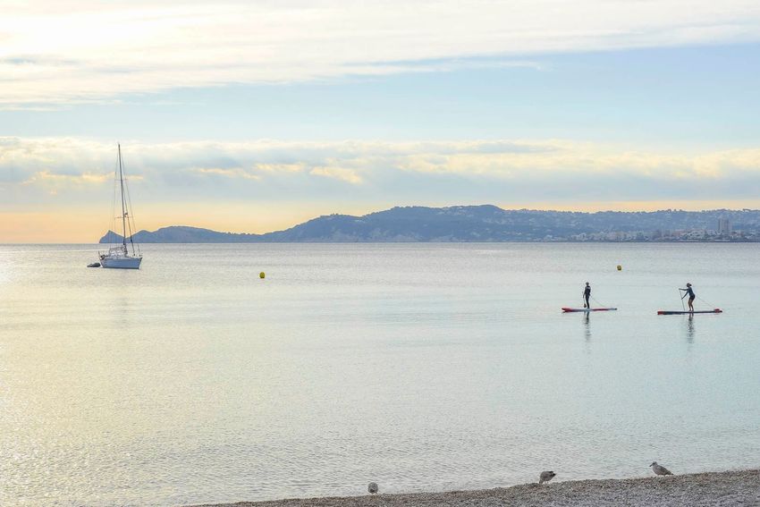 Serena escena marina con velero anclado, dos personas en paddleboard y una costa lejana bajo cielo nublado. Orilla de guijarros en primer plano. Serena escena marina con velero anclado, dos personas en paddleboard y una costa lejana bajo cielo nublado. Orilla de guijarros en primer plano.