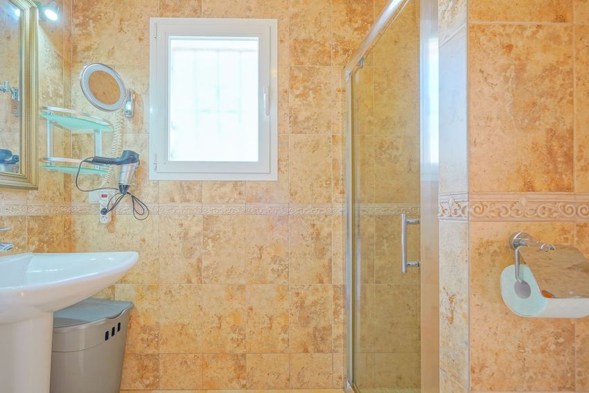 Baño revestido con azulejos de tonos tierra y cenefa decorativa. Incluye lavabo de pedestal, espejo con iluminación y cabina de ducha de cristal, junto a una ventana.