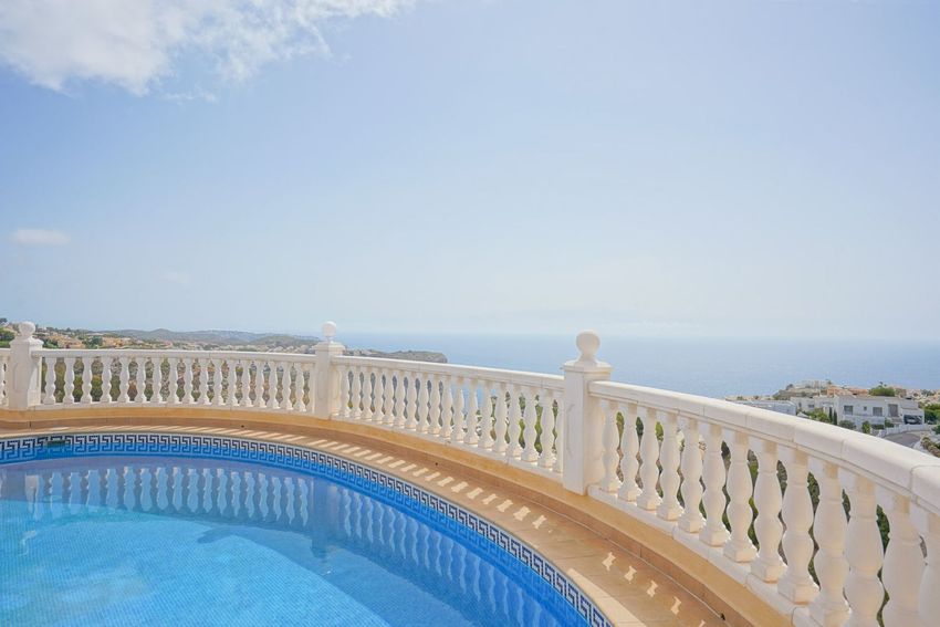 Piscina curva con mosaico azul y borde de patrón griego, enmarcada por una balustrada blanca que ofrece vistas al mar.
