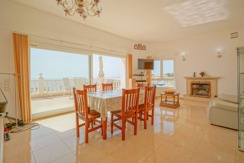 Salón-comedor luminoso con acceso directo a una terraza con barandilla blanca y vistas al mar, complementado por una chimenea de piedra y mesa de madera.