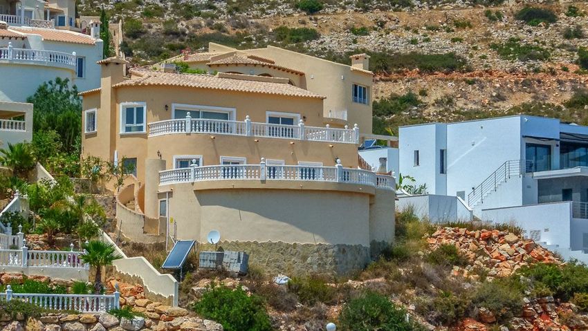 Villa mediterránea de varios niveles con fachada color arena y tejado de terracota, balcones curvos con balaustradas blancas, integrada en ladera aterrazada con paneles solares.