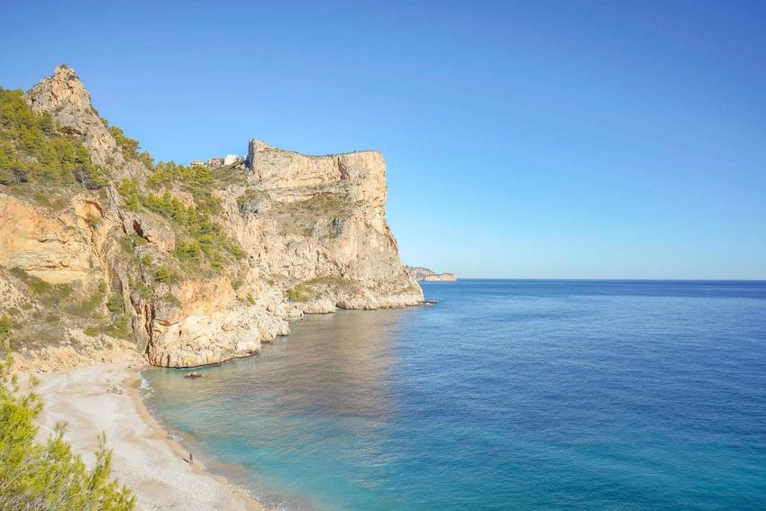 Vista panorámica de la Cala Moraig con su playa de guijarros, aguas turquesas y los imponentes acantilados de la Cumbre del Sol bajo un cielo azul claro.