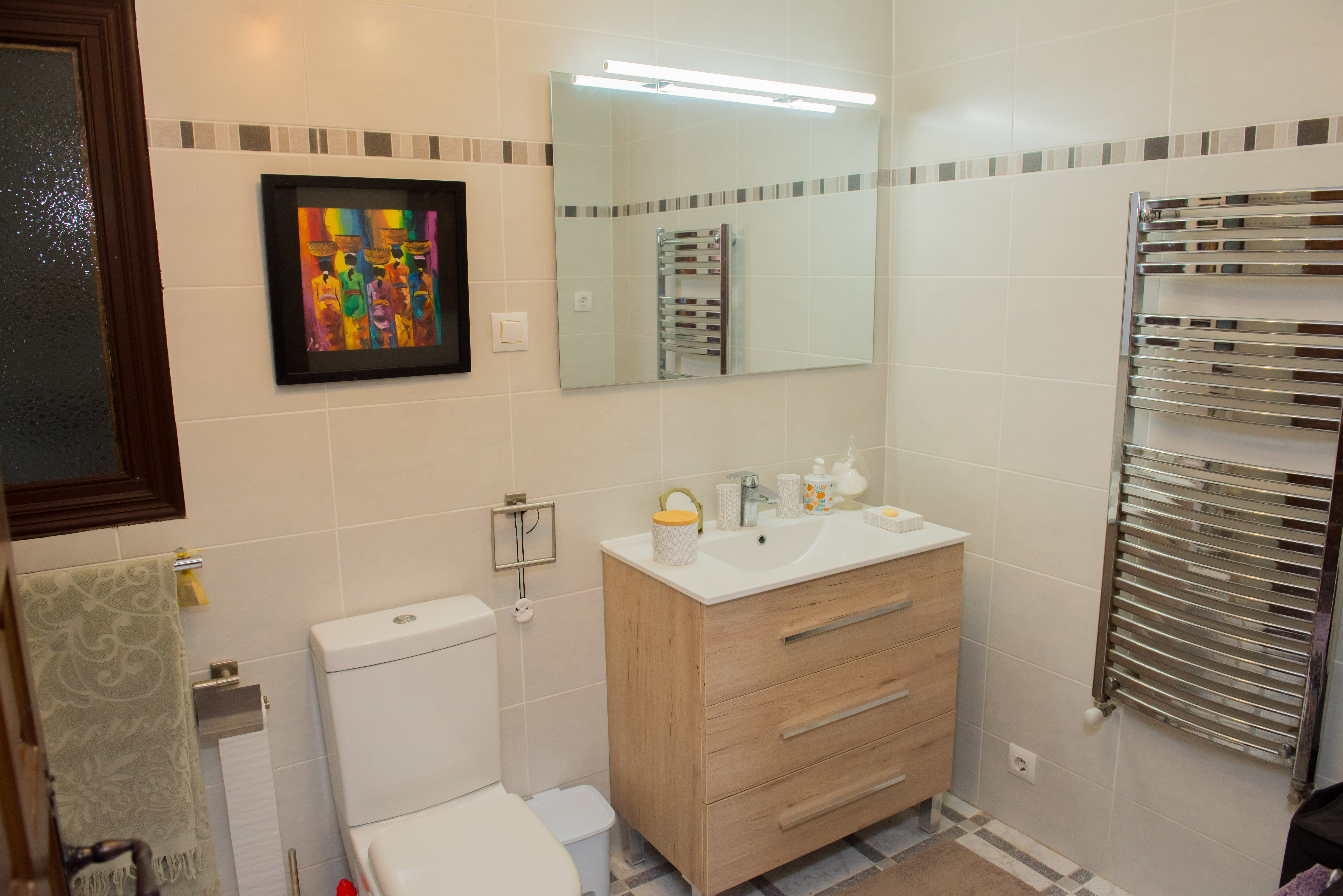 Cuarto de baño con azulejos color crema, tocador de madera clara y toallero térmico cromado.