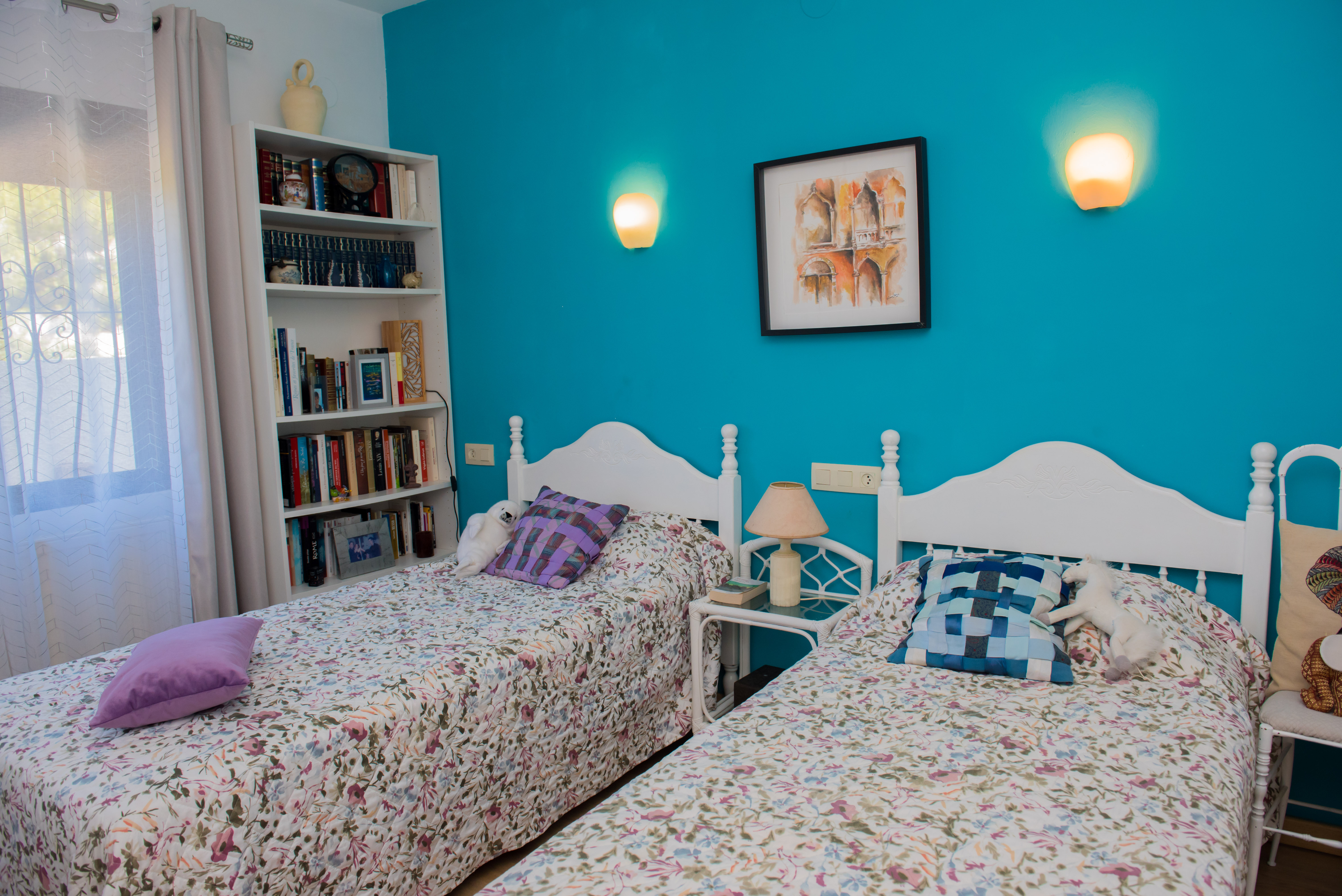 Habitación con dos camas individuales, pared azul turquesa y estantería blanca con libros.