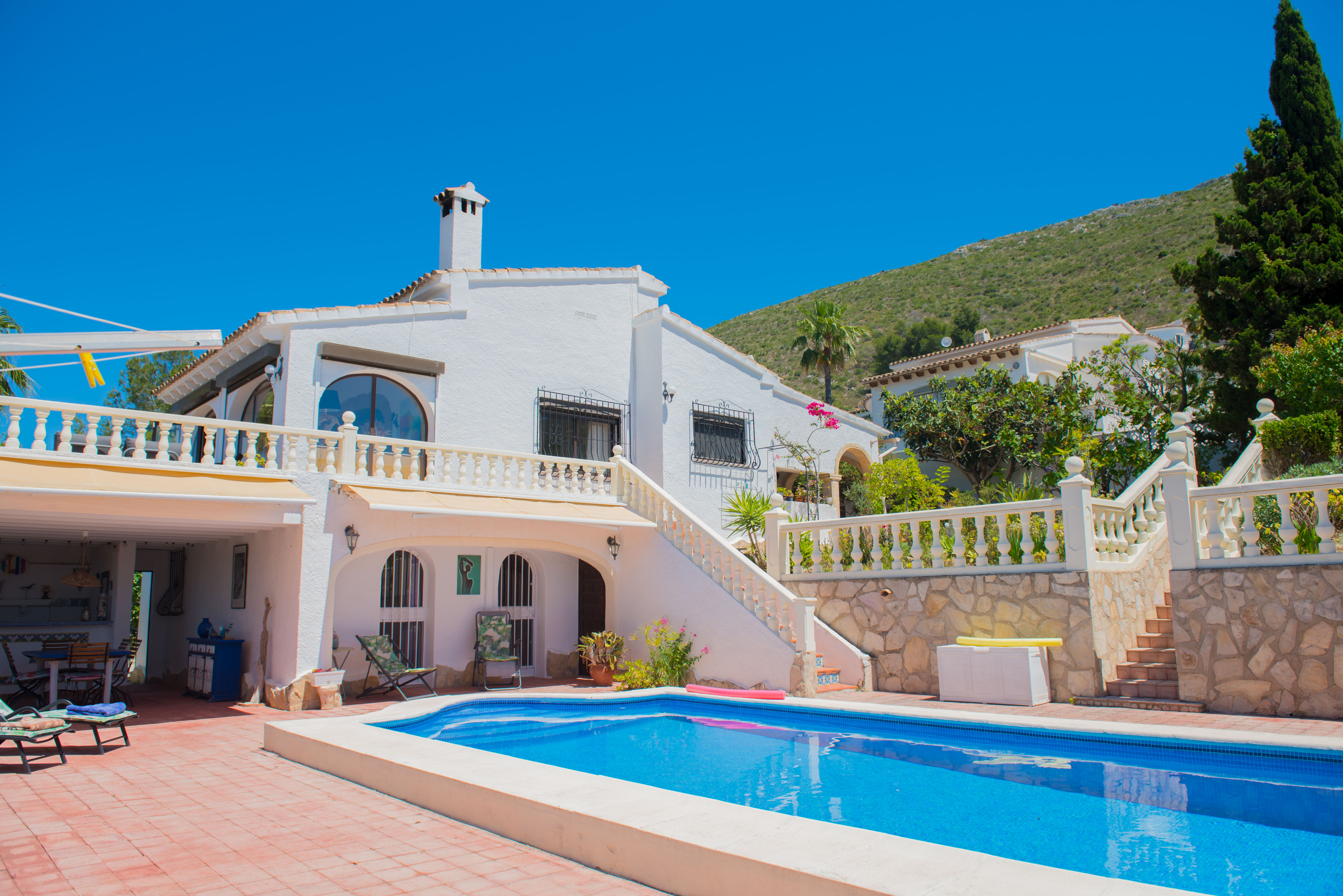 Vista de la piscina con baldosas rojas y casa blanca de estilo mediterráneo con escalera de piedra.