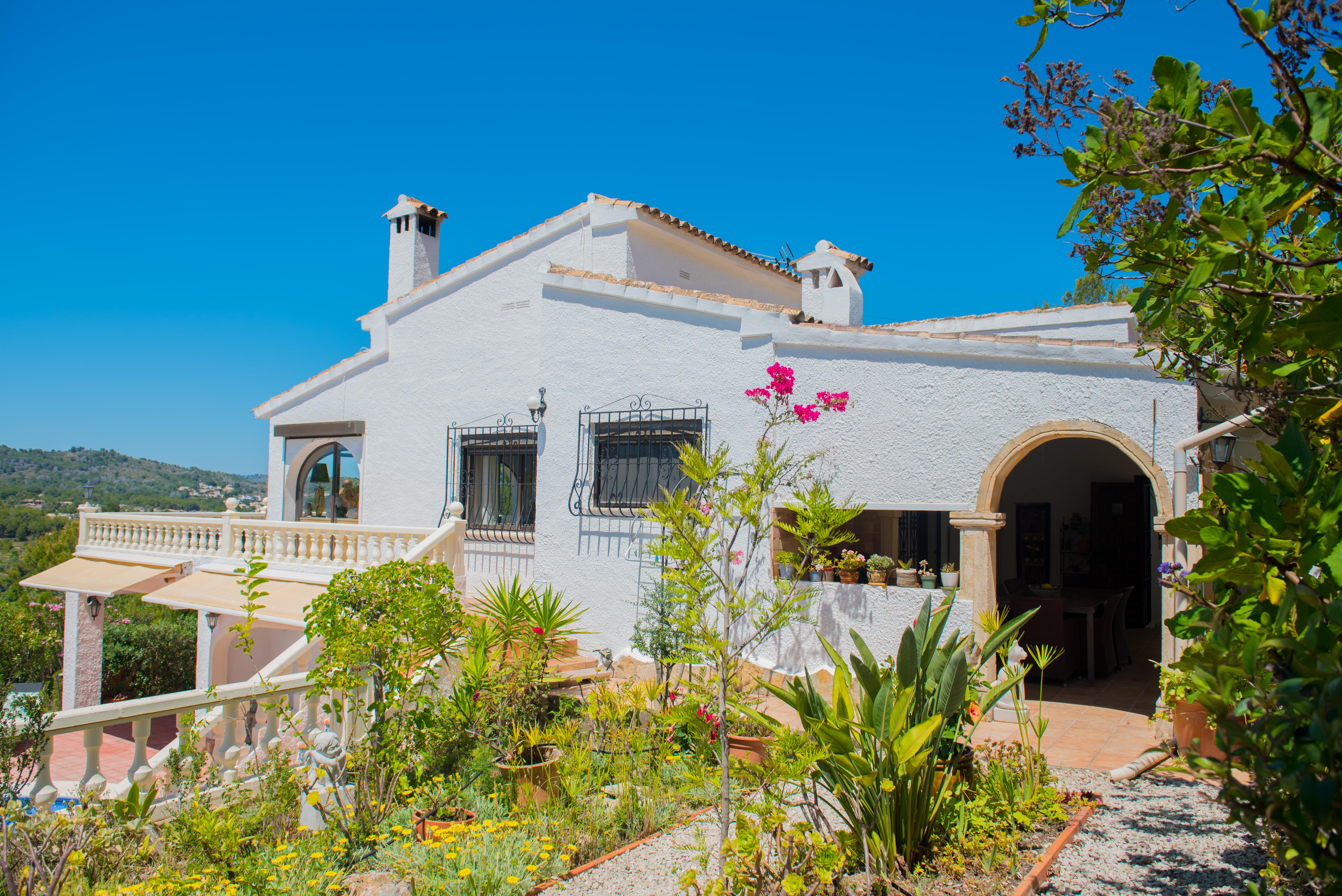 Vista exterior de la villa mediterránea con jardín, paredes blancas texturizadas y balcones con balaustrada.