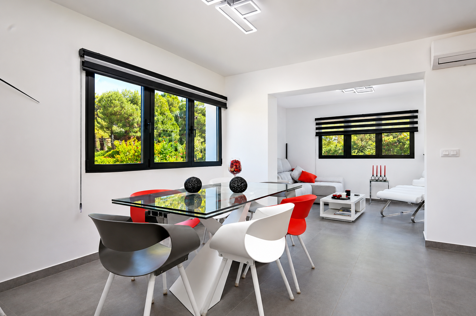 Comedor luminoso con mesa de cristal, sillas de diseño y vistas al jardín a través de ventanas con persianas.