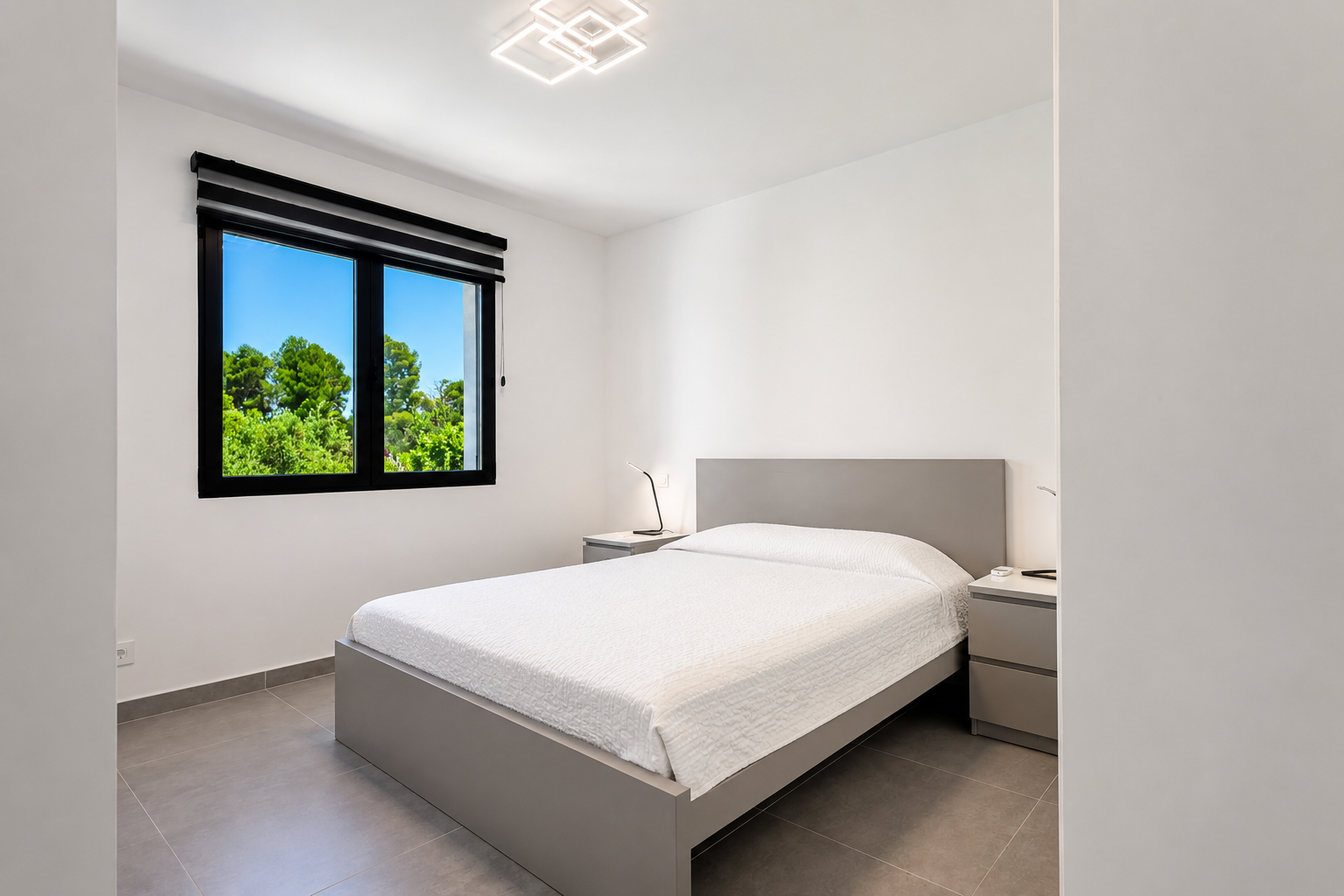 Dormitorio luminoso con cama doble gris, luz moderna y vistas al jardín a través de la ventana con marco negro.