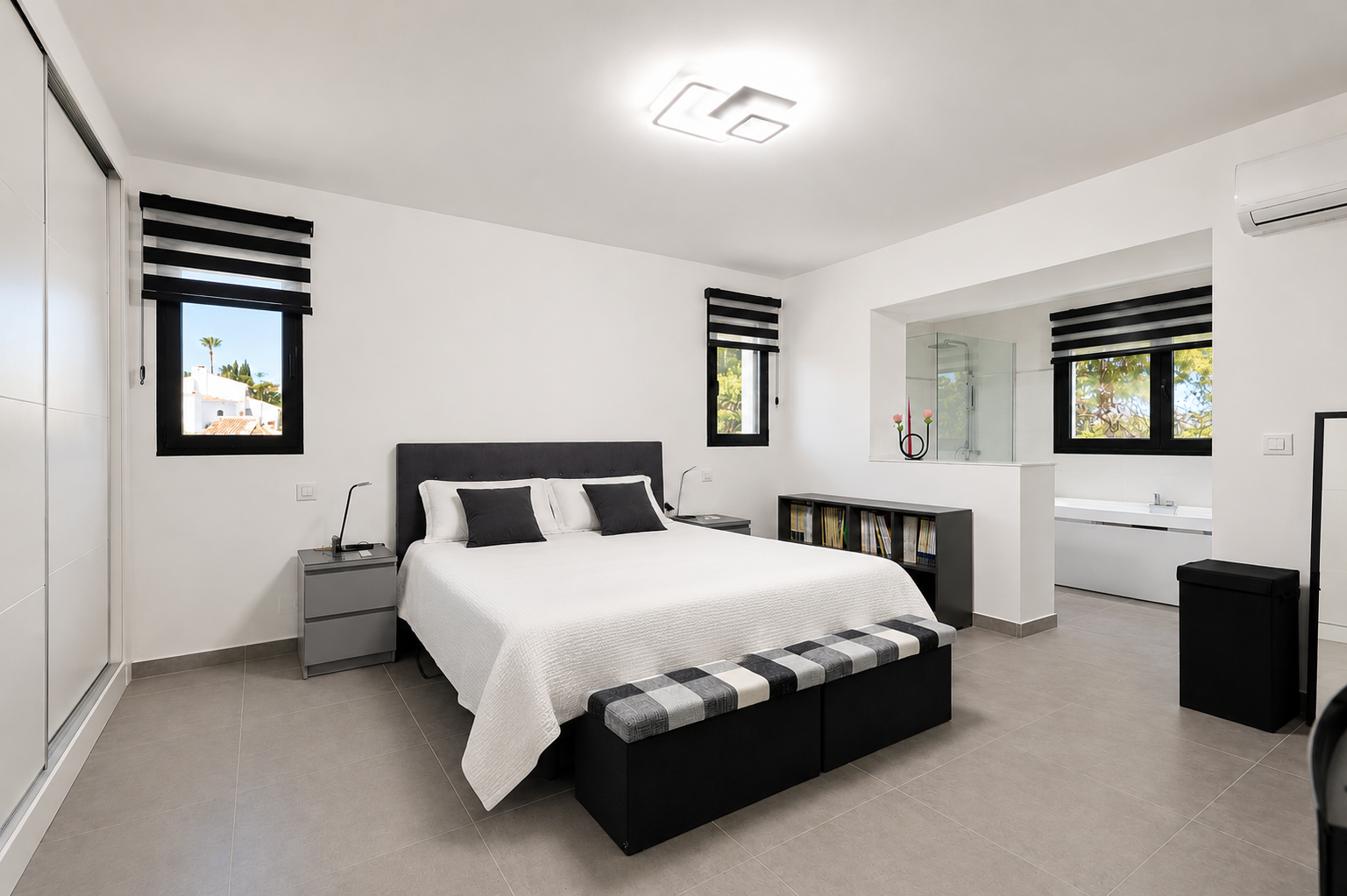 Luminoso dormitorio con suelo de baldosas grises, cama doble con cabecero tapizado y baño adyacente a través de una abertura.