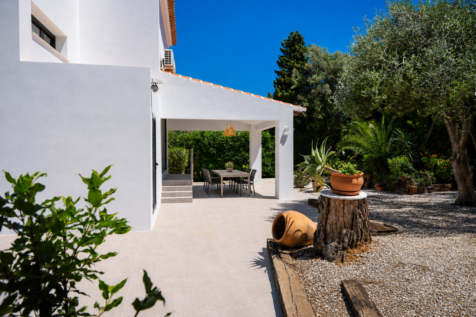 Terraza cubierta con comedor exterior, rodeada de jardín mediterráneo y gravilla blanca.