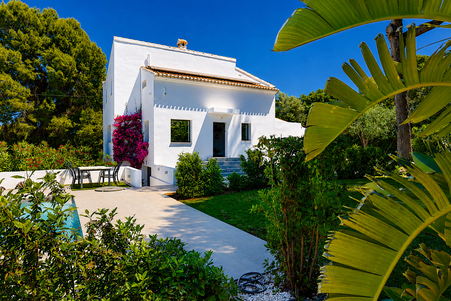 Vista exterior de la villa blanca con jardín, piscina y buganvillas en la fachada.