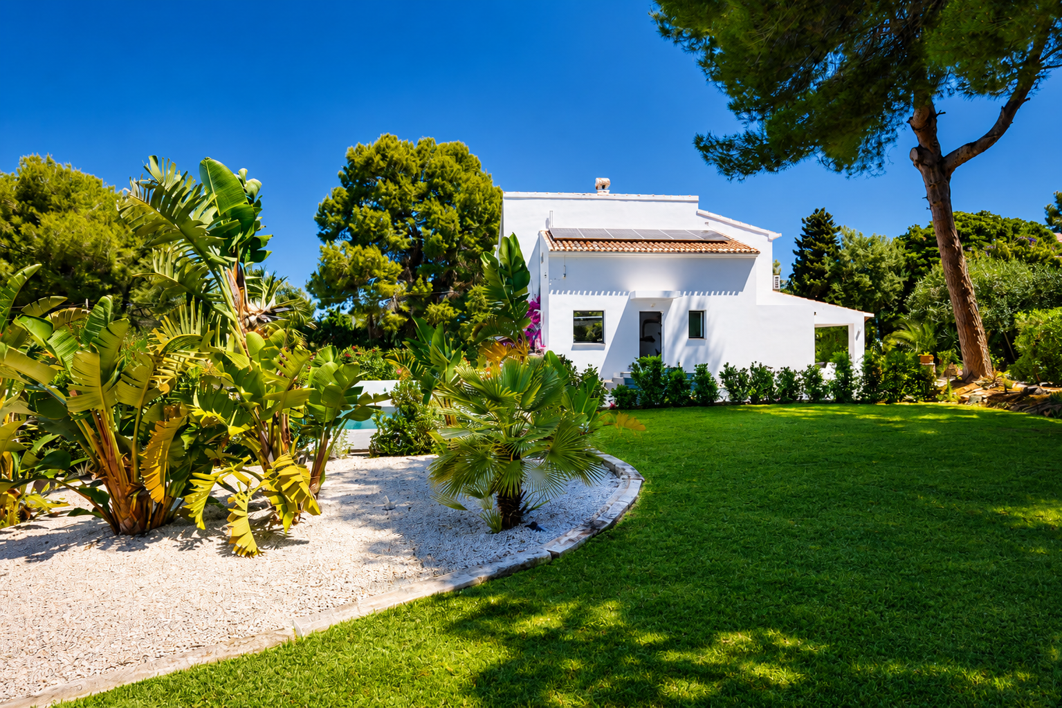 Vista exterior de la villa con fachada blanca, jardín con césped y vegetación tropical.