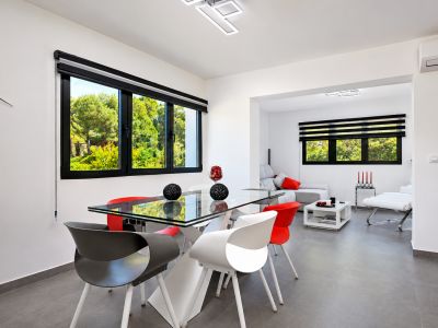 Comedor luminoso con mesa de cristal, sillas de diseño y vistas al jardín a través de ventanas con persianas.