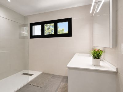 Cuarto de baño moderno con ducha de vidrio, lavabo blanco y ventana con marco negro que ilumina el espacio.