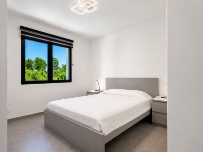 Dormitorio luminoso con cama doble gris, luz moderna y vistas al jardín a través de la ventana con marco negro.