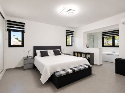 Luminoso dormitorio con suelo de baldosas grises, cama doble con cabecero tapizado y baño adyacente a través de una abertura.