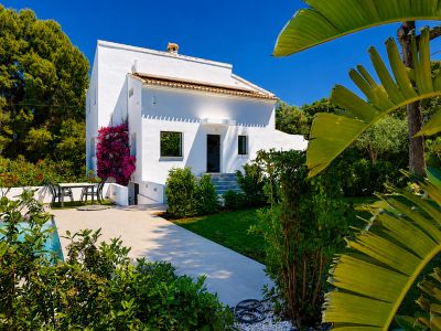 Vista exterior de la villa blanca con jardín, piscina y buganvillas en la fachada.