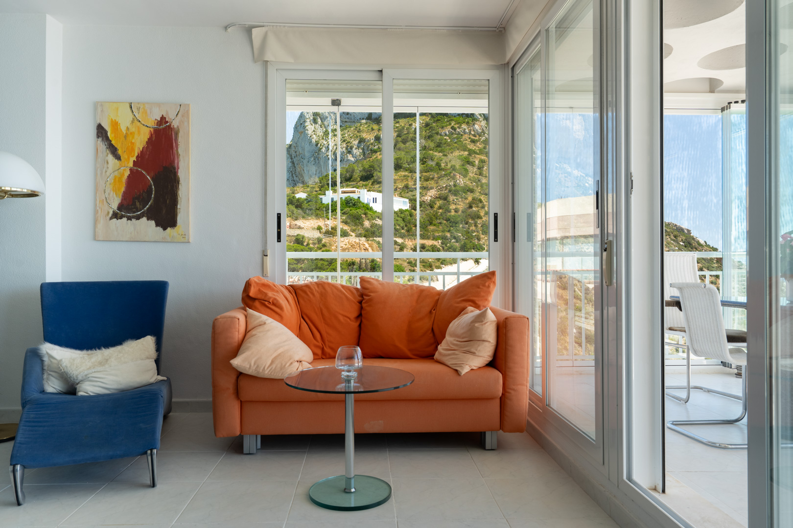 Acogedora sala de estar con sofá naranja, sillón azul y arte abstracto. Grandes ventanales enmarcan vistas a la ladera verde, con puertas correderas de cristal a una luminosa terraza cubierta.
