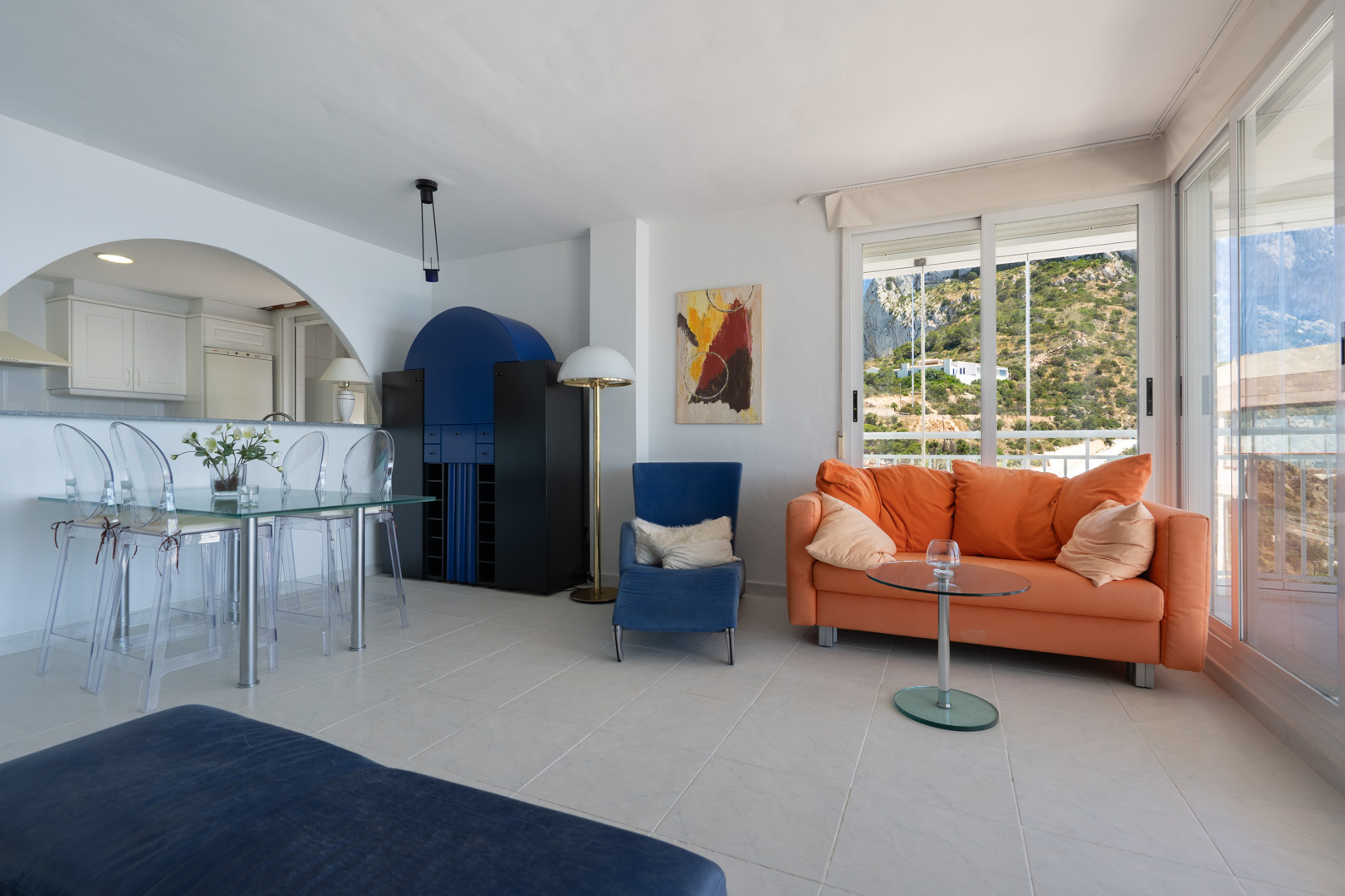 Amplio salón-comedor con sofá naranja, mesa de cristal y sillas acrílicas, junto a un mueble azul distintivo. Grandes puertas correderas ofrecen vistas a la ladera.