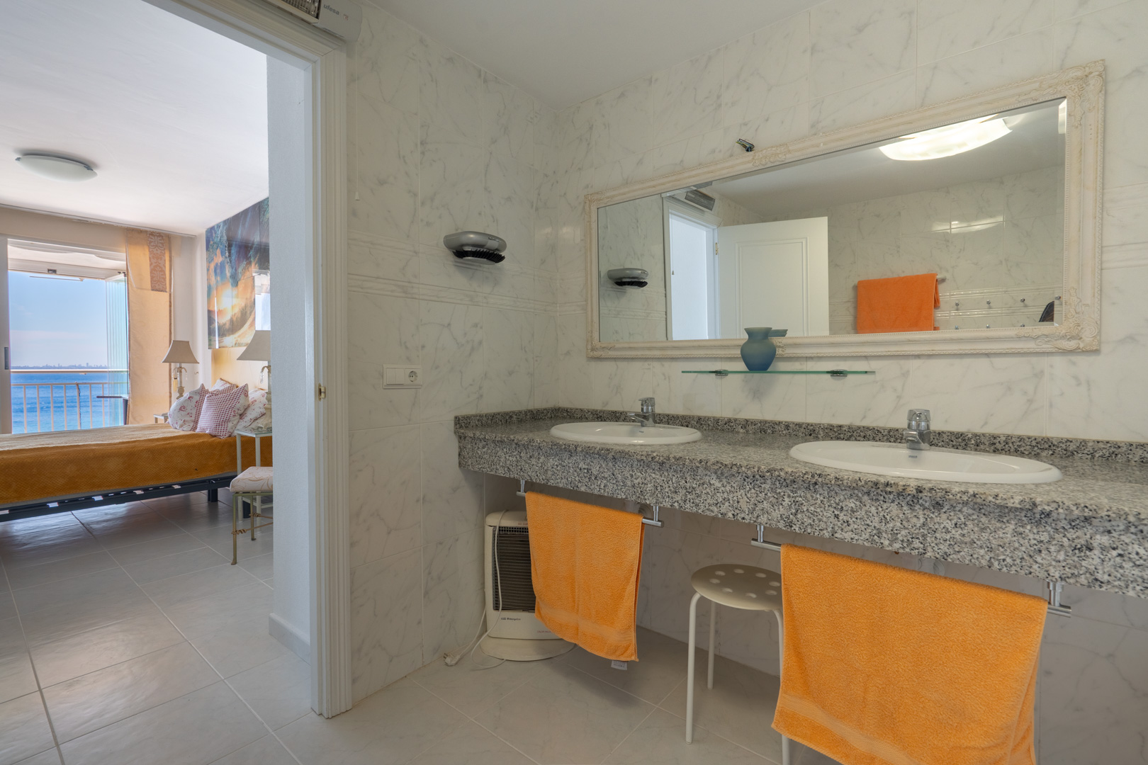Baño en suite con doble lavabo de granito y azulejos de mármol blanco. A través de la puerta abierta, se vislumbra el dormitorio adyacente con vistas al mar.