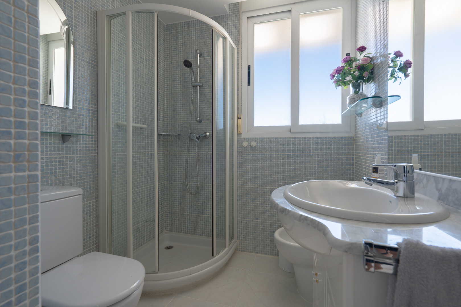Baño luminoso con azulejos de mosaico azul claro, cabina de ducha curva, inodoro, bidé y tocador de mármol con lavabo integrado, realzado por una gran ventana.