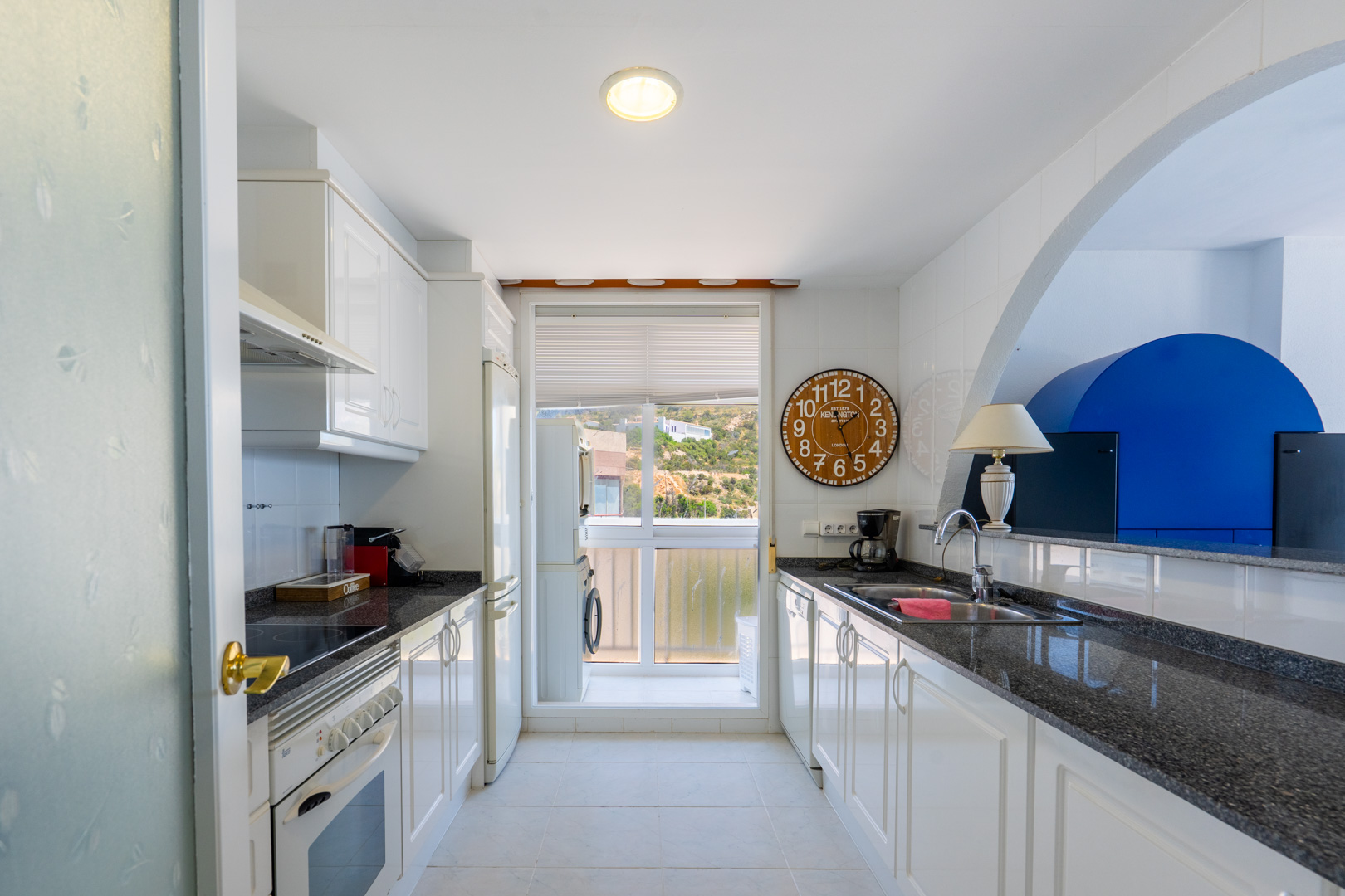 Cocina semiabierta equipada con mobiliario blanco, encimeras de granito oscuro y ventana con vistas a la ladera. Incluye zona de lavandería integrada.