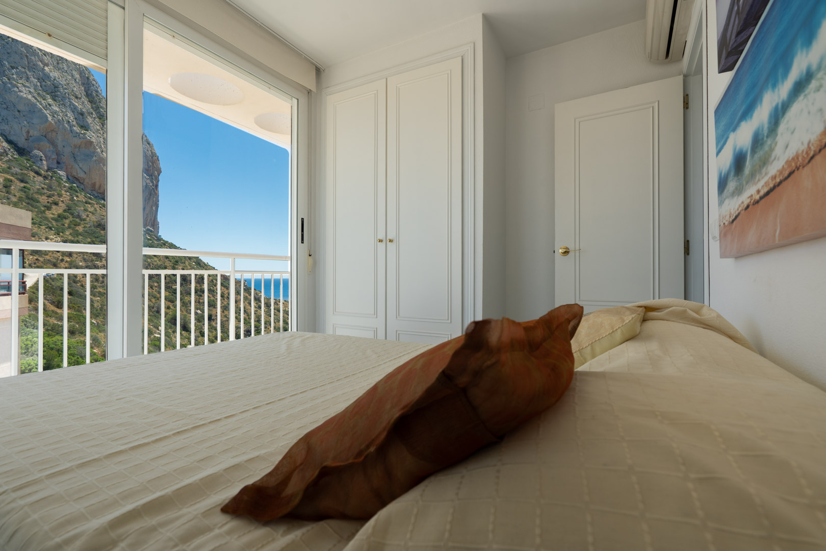 Dormitorio con armarios empotrados blancos y puertas correderas de cristal que dan a un balcón privado con vistas al Peñón de Ifach y al mar.