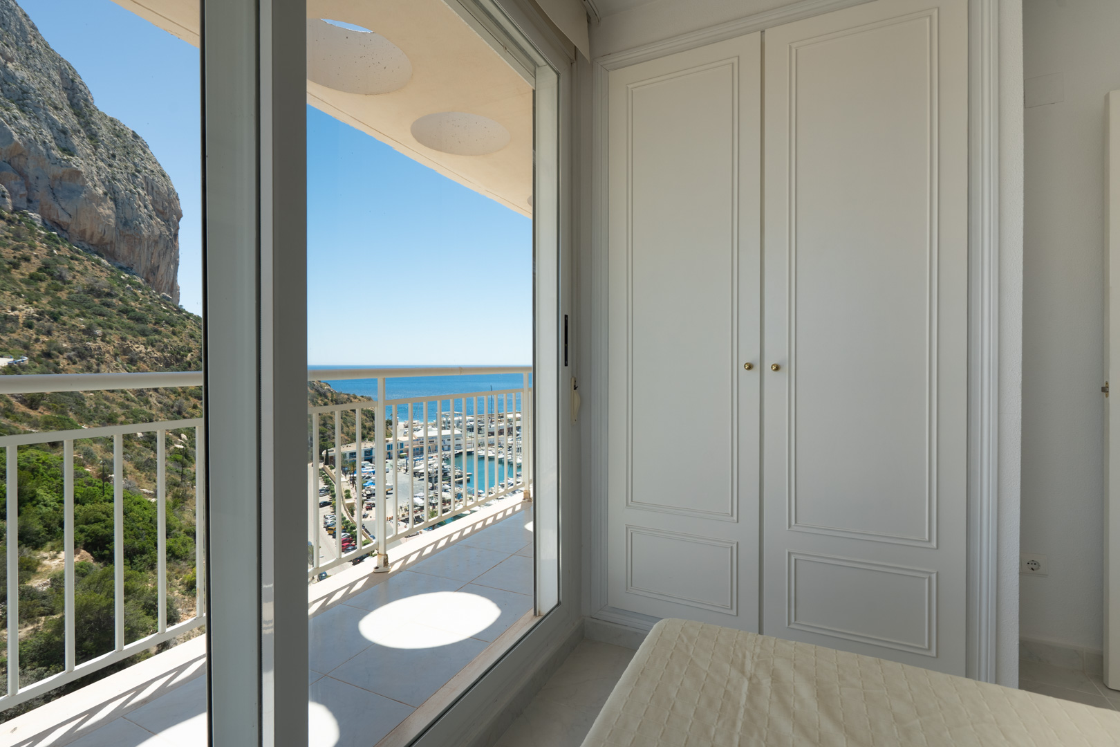 Dormitorio con elegante armario empotrado blanco y acceso directo a balcón privado. Vistas panorámicas al puerto deportivo, el mar Mediterráneo y el icónico Peñón de Ifach.