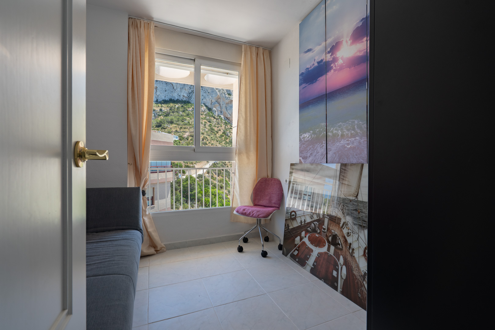 Esta versátil estancia cuenta con una ventana que ofrece vistas directas a la ladera verde, equipada con un sofá cama gris y una silla de oficina rosa. Arte mural decorativo añade un toque costero.