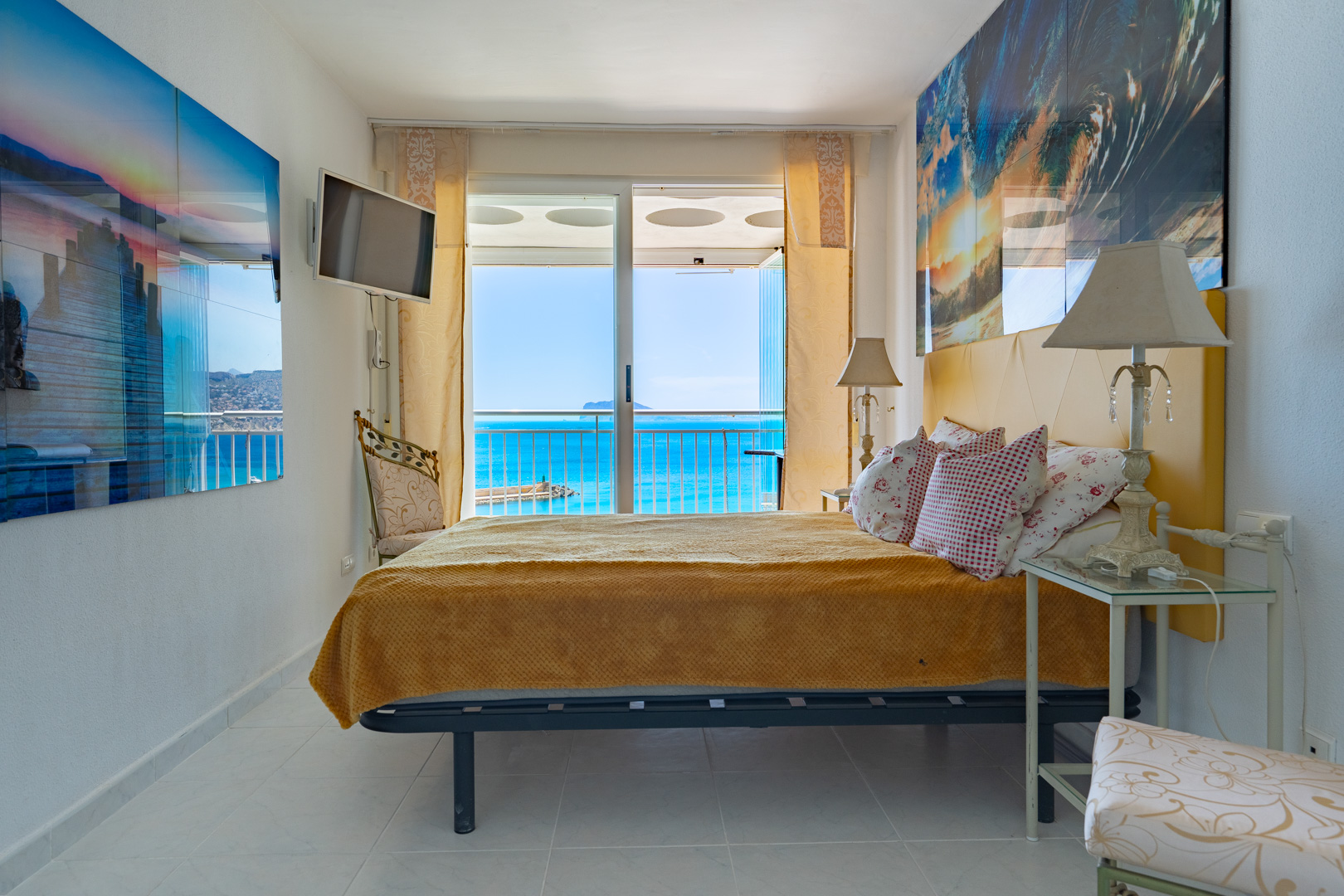 Luminoso dormitorio con cabecero amarillo y acceso directo a terraza mediante puertas correderas de cristal, ofreciendo vistas al mar azul y una masa de tierra distante.