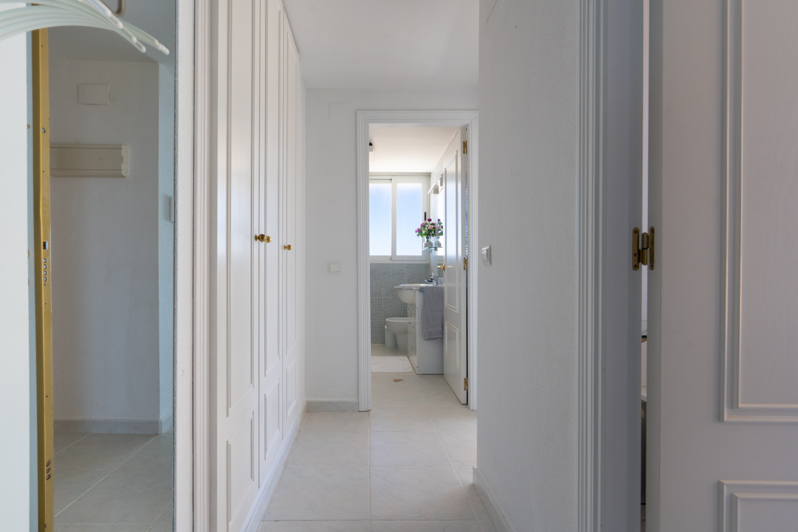 Luminoso pasillo con armarios empotrados blancos y tiradores dorados. El suelo de baldosas conduce a un baño visible a través de una puerta abierta, mostrando una ventana y azulejos de mosaico azul.