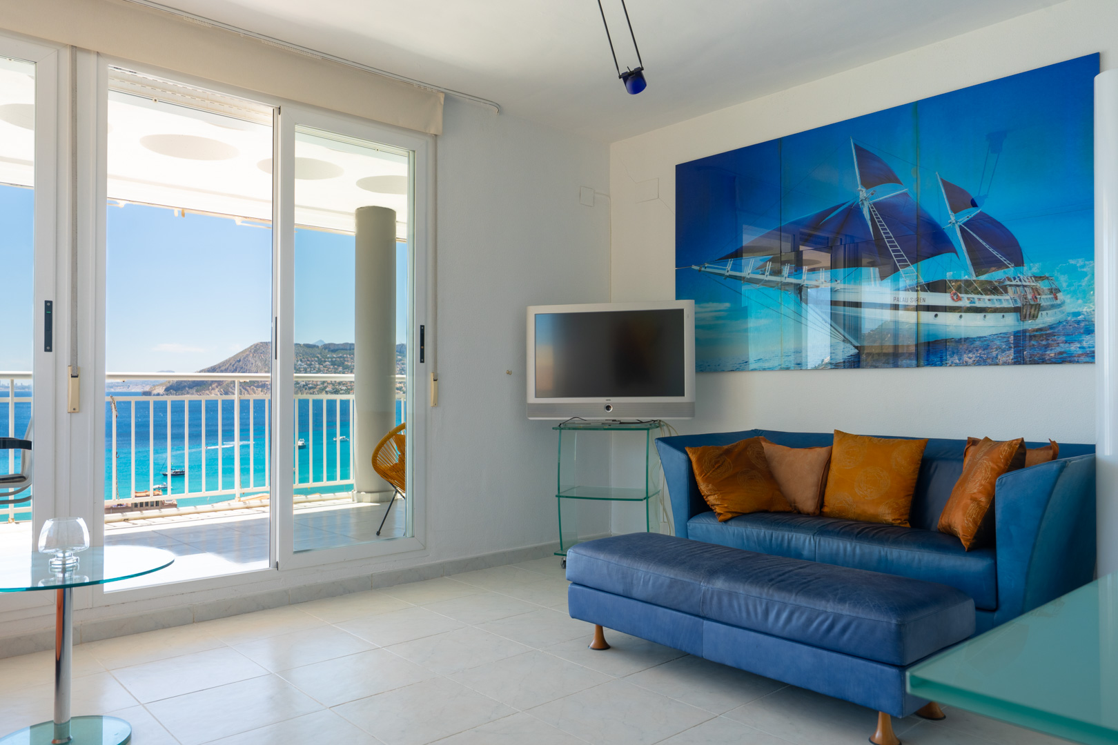 Salón luminoso con sofá azul, obra de arte de velero y puertas correderas que dan a una terraza con vistas panorámicas al mar y la costa.