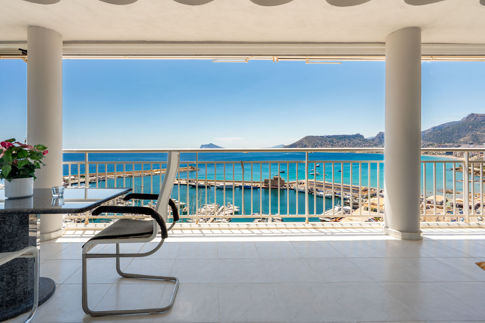 Terraza cubierta con suelo de baldosas blancas y columnas estructurales, ofreciendo vistas panorámicas directas al puerto deportivo de Calpe, el mar Mediterráneo y montañas lejanas.
