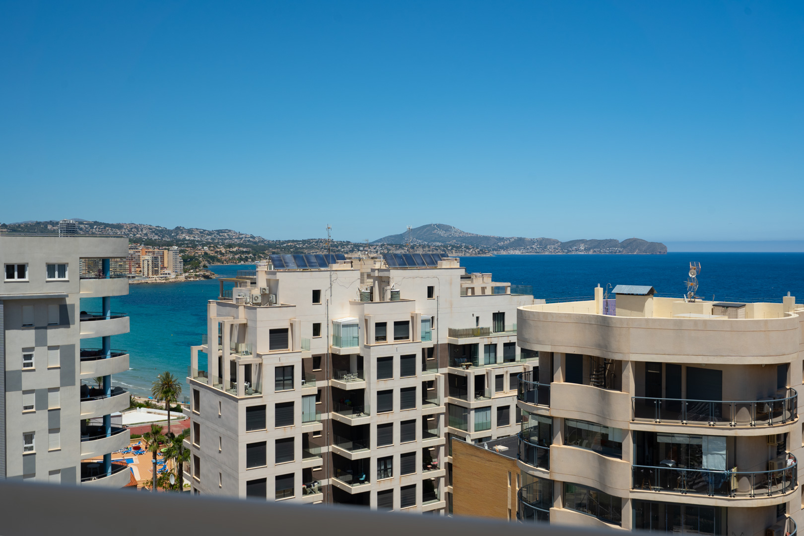 Vista elevada al mar desde una terraza, mostrando arquitectura costera moderna, el vibrante Mediterráneo y montañas lejanas. Piscina comunitaria y palmeras visibles.
