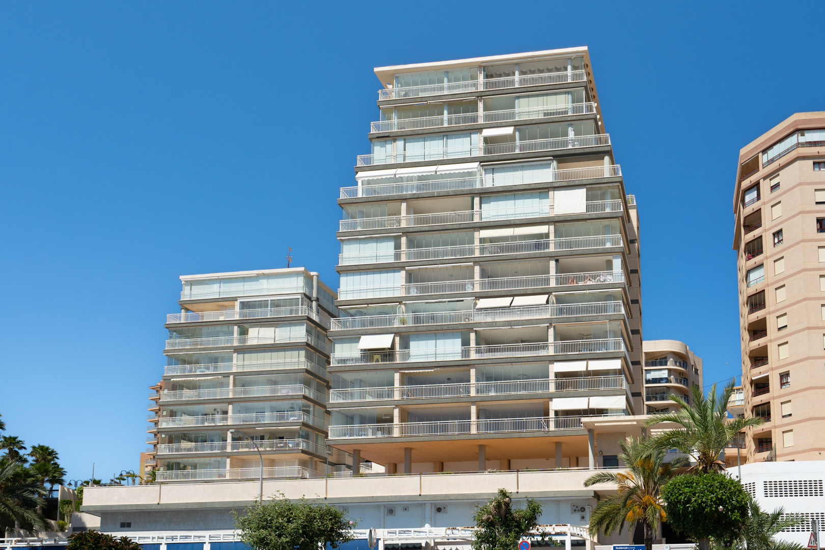 Vista exterior de un moderno edificio residencial de varias plantas con numerosos balcones, muchos cerrados con cortinas de cristal y barandillas blancas, bajo un cielo azul claro. Palmeras en primer plano.