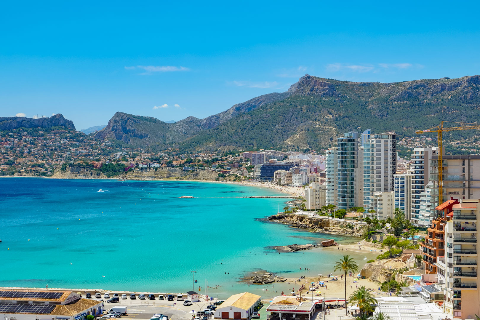 Vista panorámica de la bahía de Calpe con aguas turquesas, playas de arena, modernos edificios costeros y montañas verdes bajo un cielo despejado.