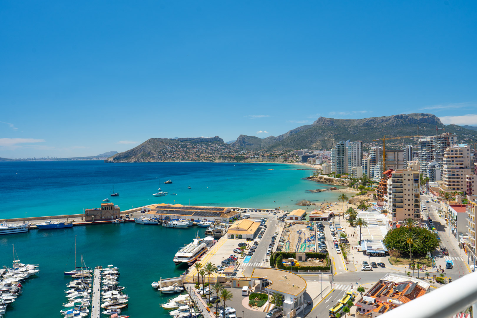 Vista panorámica del animado puerto deportivo de Calpe con numerosos yates, la bahía turquesa, la playa de arena y las montañas al fondo.