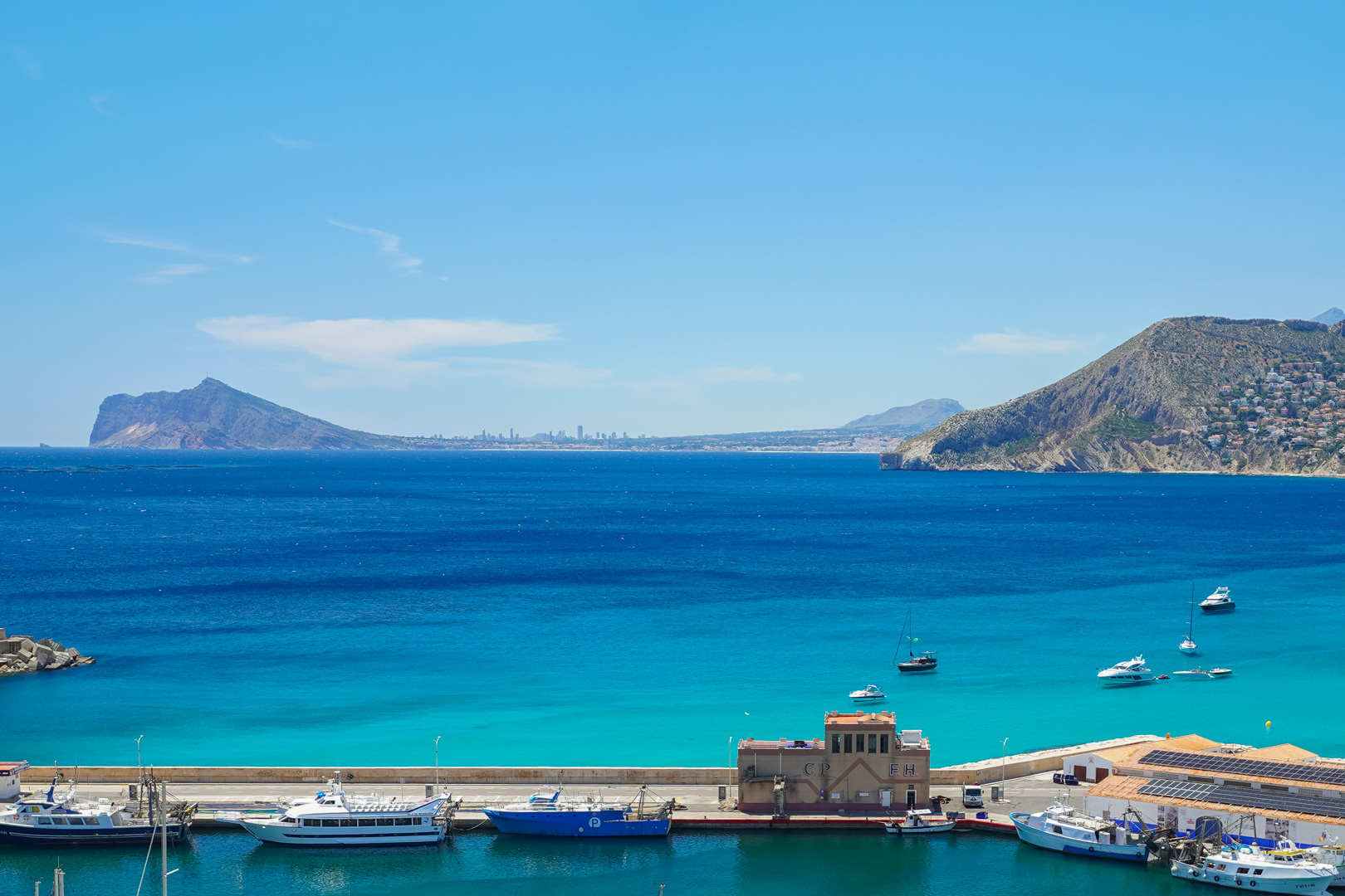 Vista panorámica del puerto deportivo de Calpe con embarcaciones, aguas turquesas y montañas lejanas. El Peñón de Ifach domina a la derecha, con un skyline urbano al fondo.