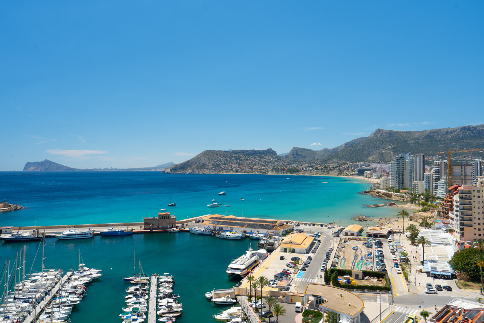 Vista panorámica del puerto deportivo de Calpe con yates, la vibrante bahía de aguas turquesas y una playa de arena junto a edificios costeros y montañas.