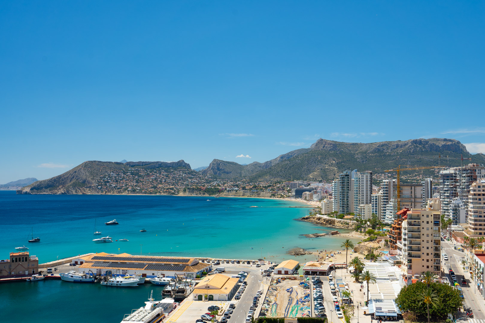 Vista panorámica del puerto deportivo de Calpe, la bahía turquesa y la Playa de La Fossa, enmarcada por montañas lejanas bajo un cielo azul claro.