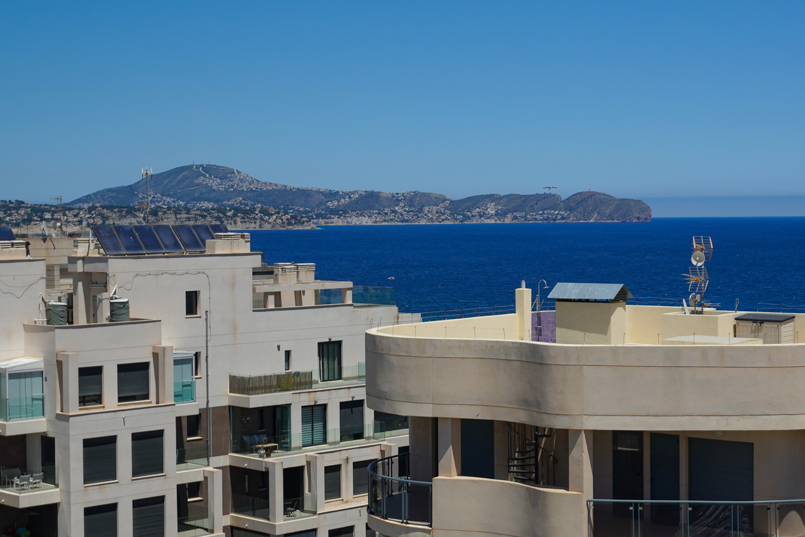 Vistas panorámicas al mar desde una planta alta, con arquitectura moderna adyacente, paneles solares y una costa montañosa urbanizada a lo lejos.