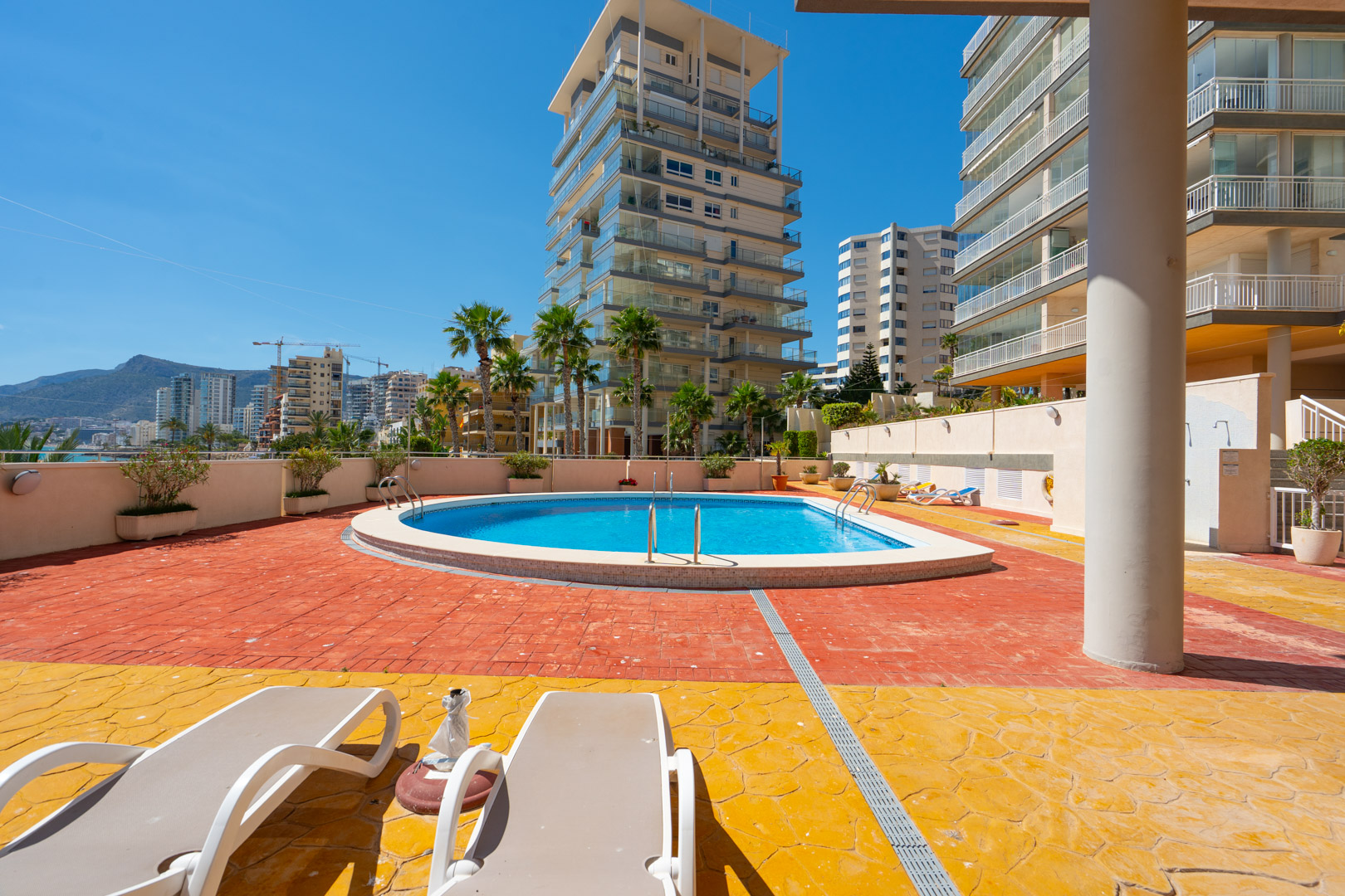 Zona de piscina comunitaria con agua azul cristalina, tumbonas y pavimento bicolor, enmarcada por edificios modernos y palmeras bajo un cielo despejado.